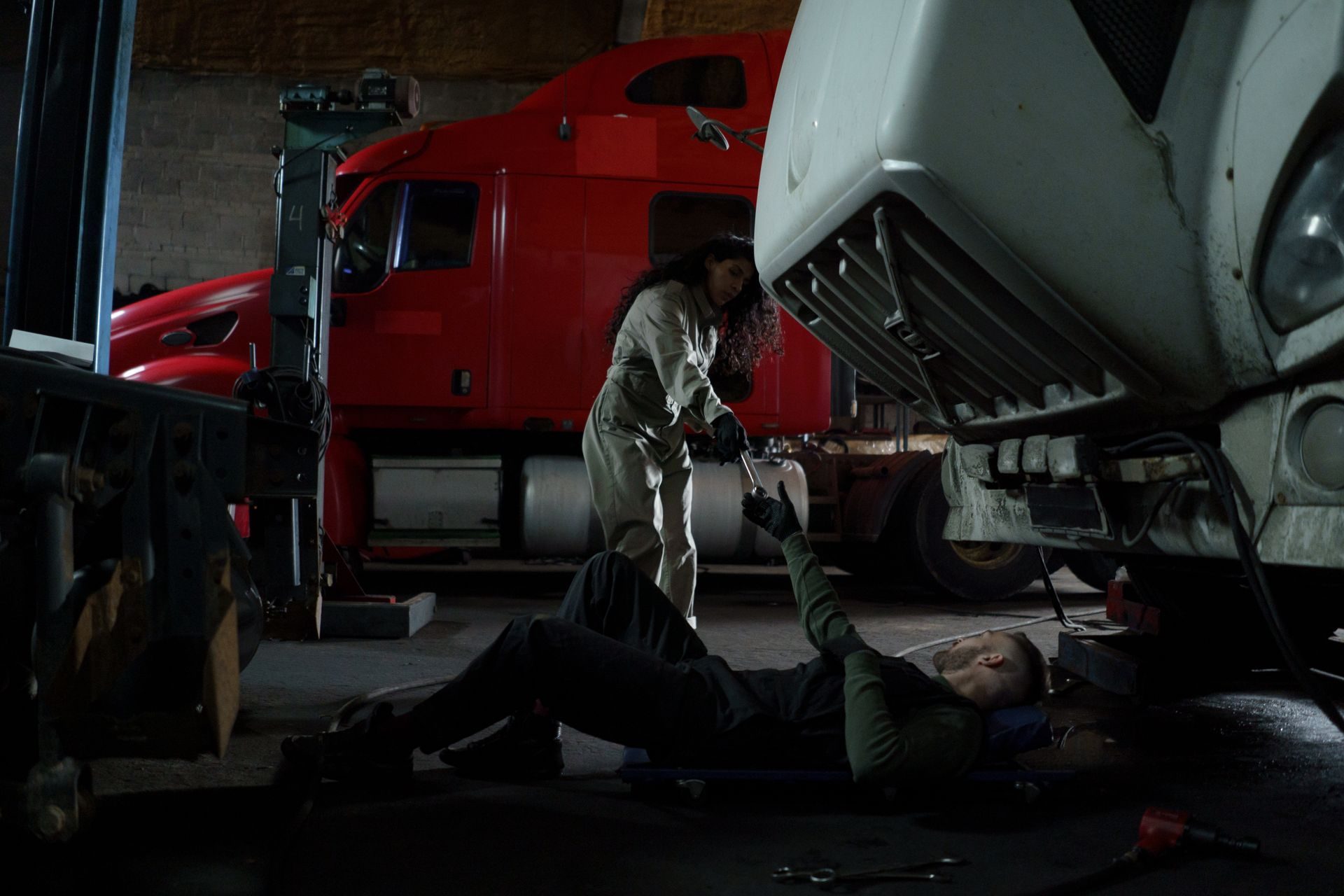 Two diesel technicians repairing a semi truck, one passing a wrench under the cab
